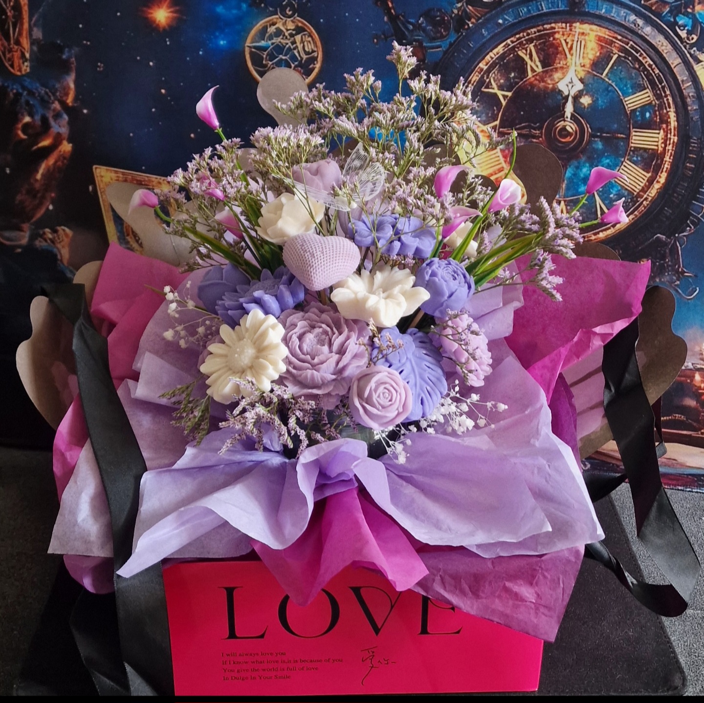 représentation d'une composition florale en bougie moulée avec feuillage et fleurs séchées dans un support carré rose avec inscription love